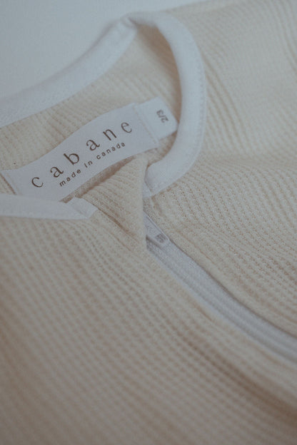 Close-up image of a beige garment with a white zipper and a white fabric tag displaying the brand name "cabane childrenswear" and the text "made in canada" in lowercase. The fabric has a textured, ribbed pattern, and the garment size is indicated as "2T" on the tag. This seasonless unisex outfit, known as THE ORB SUIT WAFFLE baby + kid, is perfect for easy diaper changes.