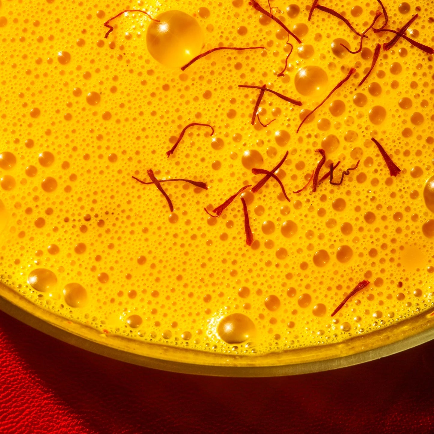 Close-up of a vibrant saffron latte with frothy bubbles on the surface, garnished with thin red saffron strands—likely a Warm Feelings® Saffron Latte by THE FULLEST, known for its caffeine-free properties and mood-improving benefits. The background appears to be a red cloth.