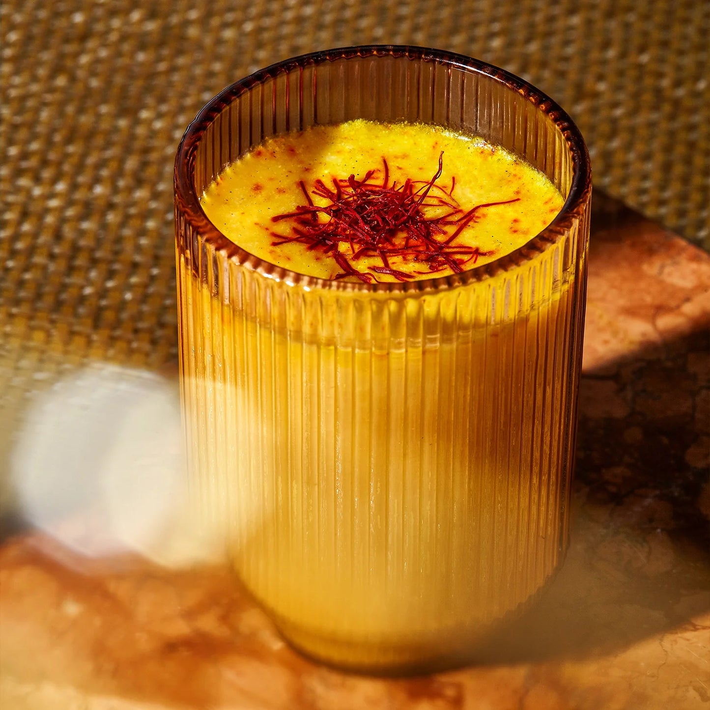 A luxurious Warm Feelings® Saffron Latte from THE FULLEST, golden-colored and caffeine-free, is served in a ribbed amber glass with saffron strands garnishing the top. It sits on a brown, marble-like surface against a backdrop featuring a textured, woven pattern. 