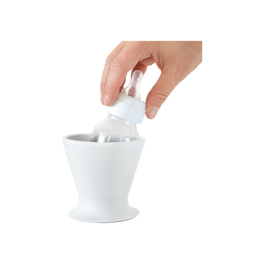 A hand places a baby bottle into the white, cone-shaped bökee Baby Bottle Holder by gripping the bottle's neck and angling it as it's lowered into the holder. Ideal for one-handed bottle prep, the bökee has a wide, stable base and a narrow opening where the bottle sits.