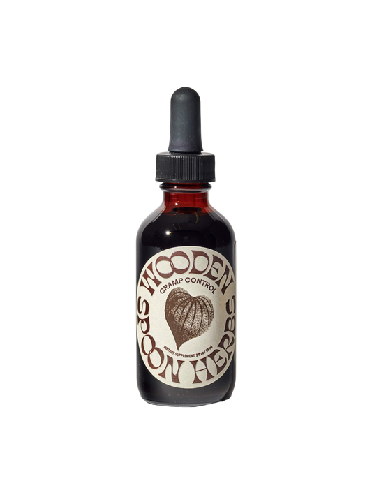 A dark glass dropper bottle labeled "Cramp Control Tincture" by WOODEN SPOON HERBS features a brown woven heart design and the text "dietary supplement 2 fl oz (59 ml)." Set against a black background, it supports a balanced cycle to help menstruate with ease.