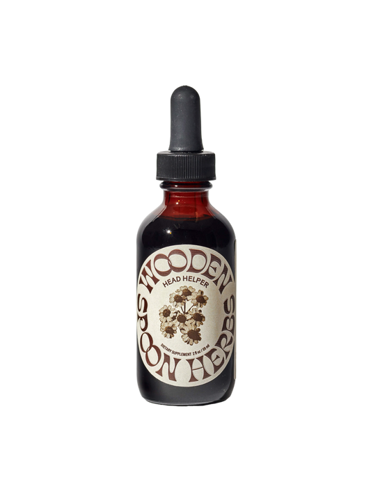 A dark amber glass bottle with a dropper lid, labeled "WOODEN SPOON HERBS Head Helper Tincture," in stylized text. The vintage-designed label features an illustration of herbs, hinting at the herbal tincture contained within.