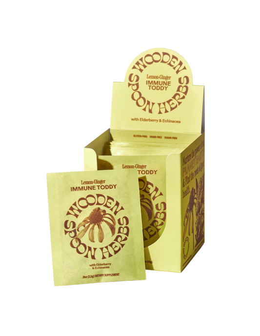 A yellow box of WOODEN SPOON HERBS Lemon-Ginger Immune Toddy Sachets, designed for immune support with elderberry and echinacea, is displayed. One sachet is positioned in front of the open box, showcasing the front label and the brown logo featuring a wooden spoon with herbs.